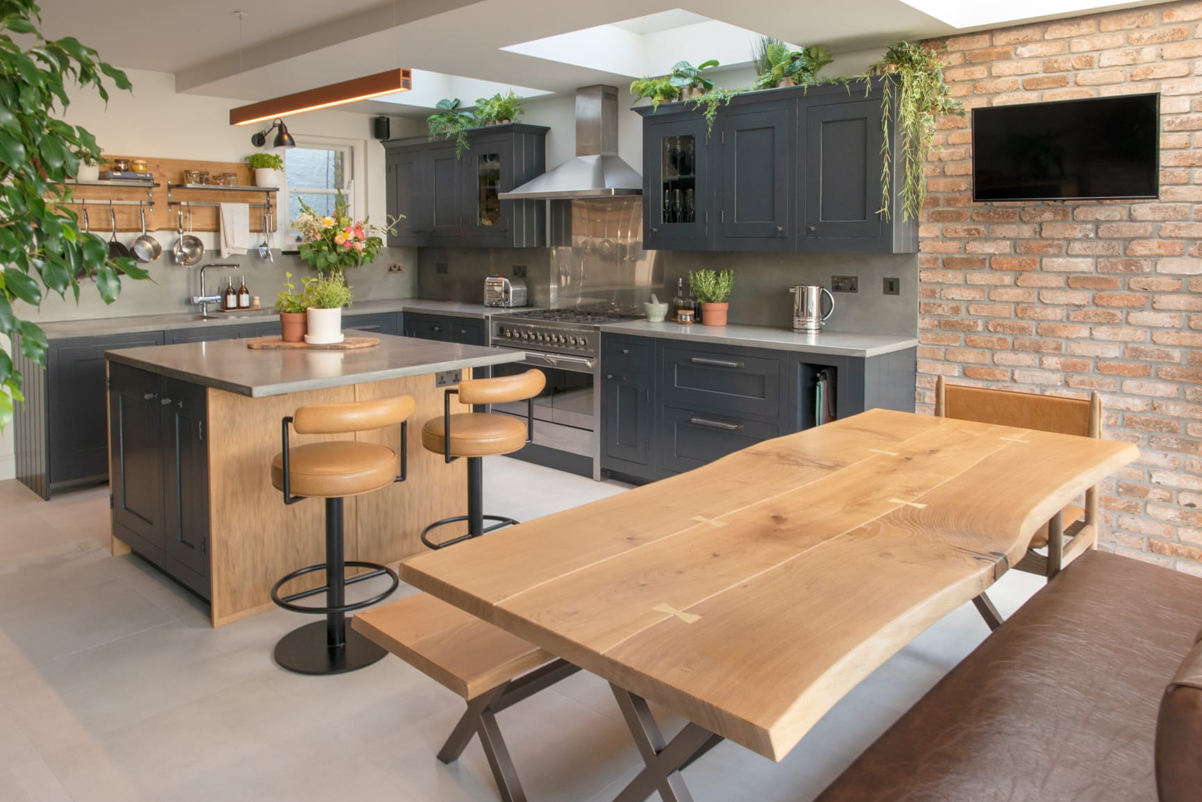 Modern kitchen with wooden table, island, barstools, plants, and exposed brick wall.