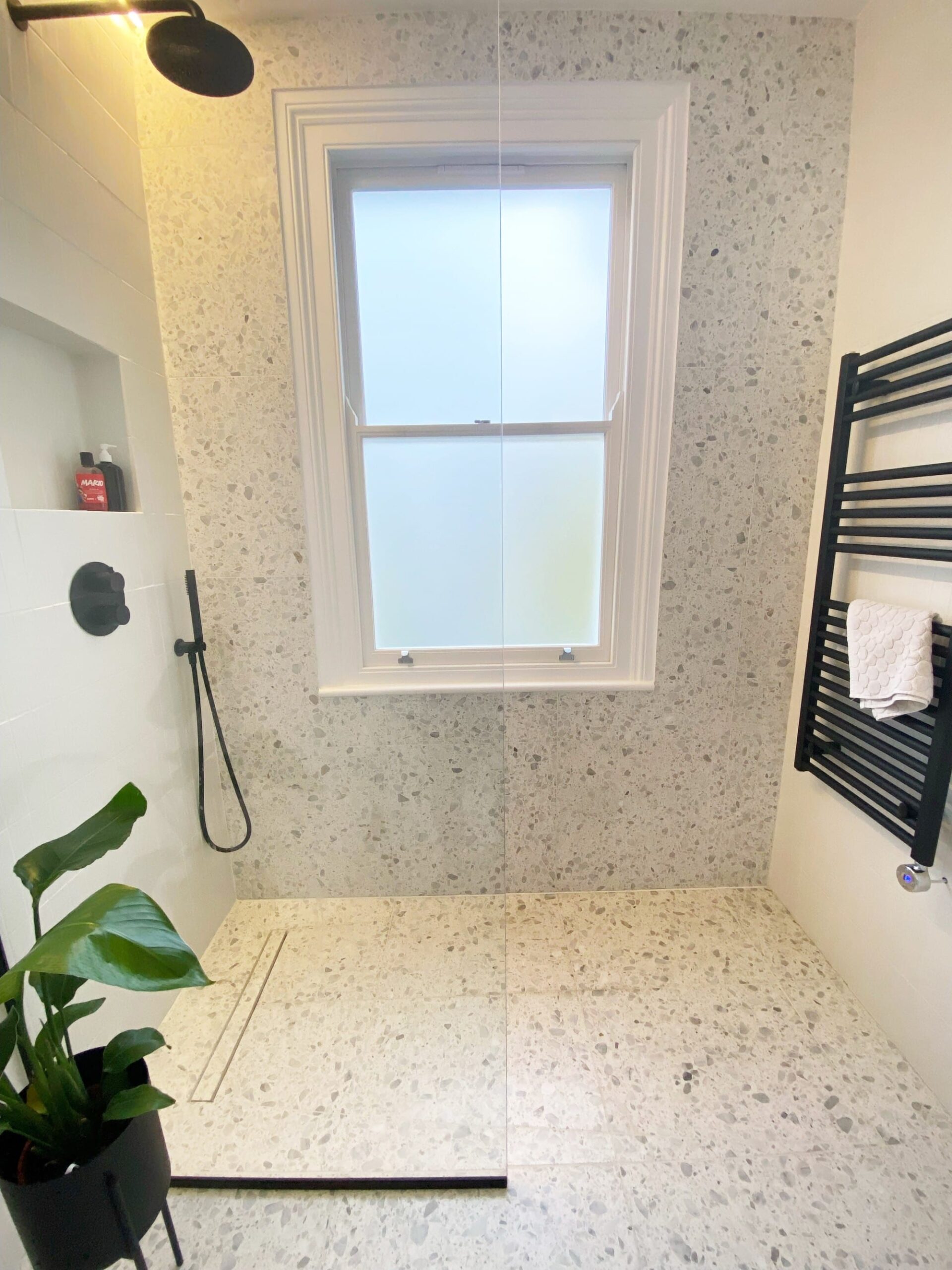 Modern bathroom with terrazzo tiles, a walk-in shower, window, towel rack, and a potted plant.