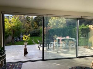 A dog runs outside through open glass doors to a sunny patio and garden.