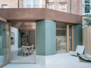 Modern house exterior with large windows, patio chairs, and a minimalist outdoor seating area.