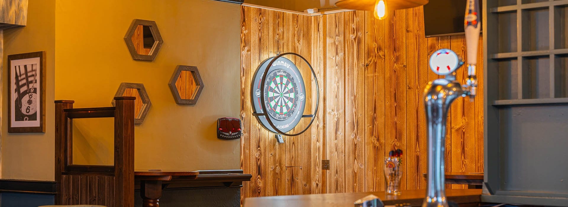 Pub interior with wood walls, a dartboard, beer tap, and framed decor visible on the wall.